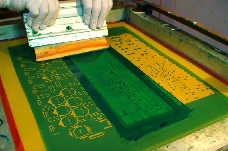 A person with white gloves operates a printing device, applying pressure to transfer green substance onto a circuit design on a PCB. The scene takes place on a workshop table, resembling a manufacturing setup for electronic components.
