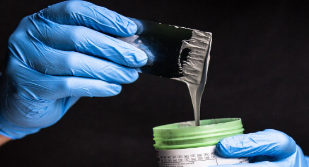 Two blue gloved hands holding spatula and plastic container for lifting the smooth grey paste.