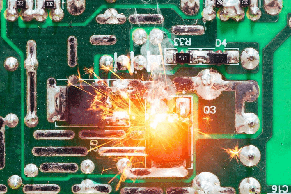 Close-up of a malfunctioning green circuit board with a bright spark at its center. Glowing red areas and silver electronic components suggest heat or damage. Out-of-focus background enhances circuit board focus. Component identifiers like