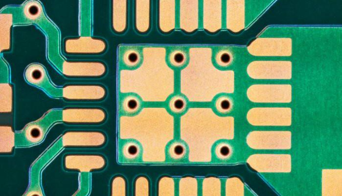 Close-up view of a green printed circuit board or microchip with a grid pattern of interlinked holes and lines. The central area stands out and features gold-plated contacts, emphasizing the precise technical design.