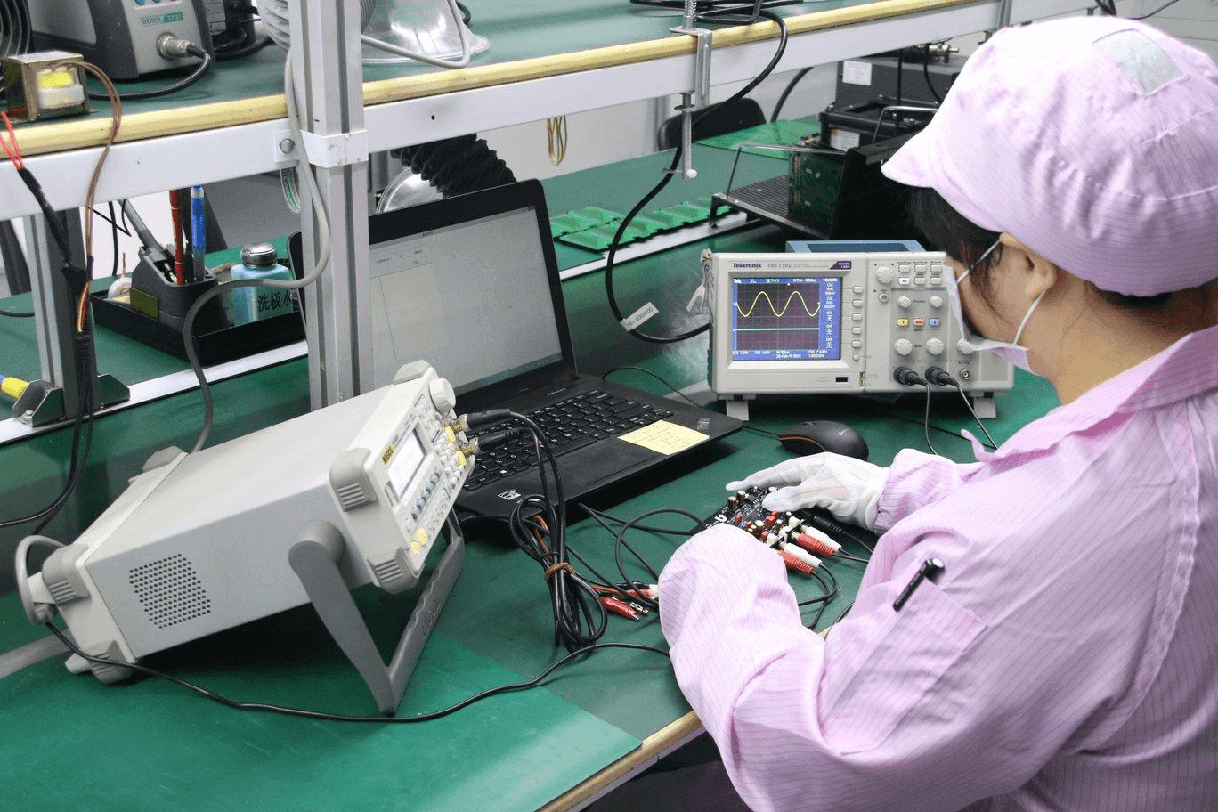 Factory worker in a pink shirt and white cap working with electronic devices. Sitting in front of a laptop connected to a circuit board with wires. Other electronic components and devices on the table. Visible clock and green-tinted surroundings, representing the factory environment.