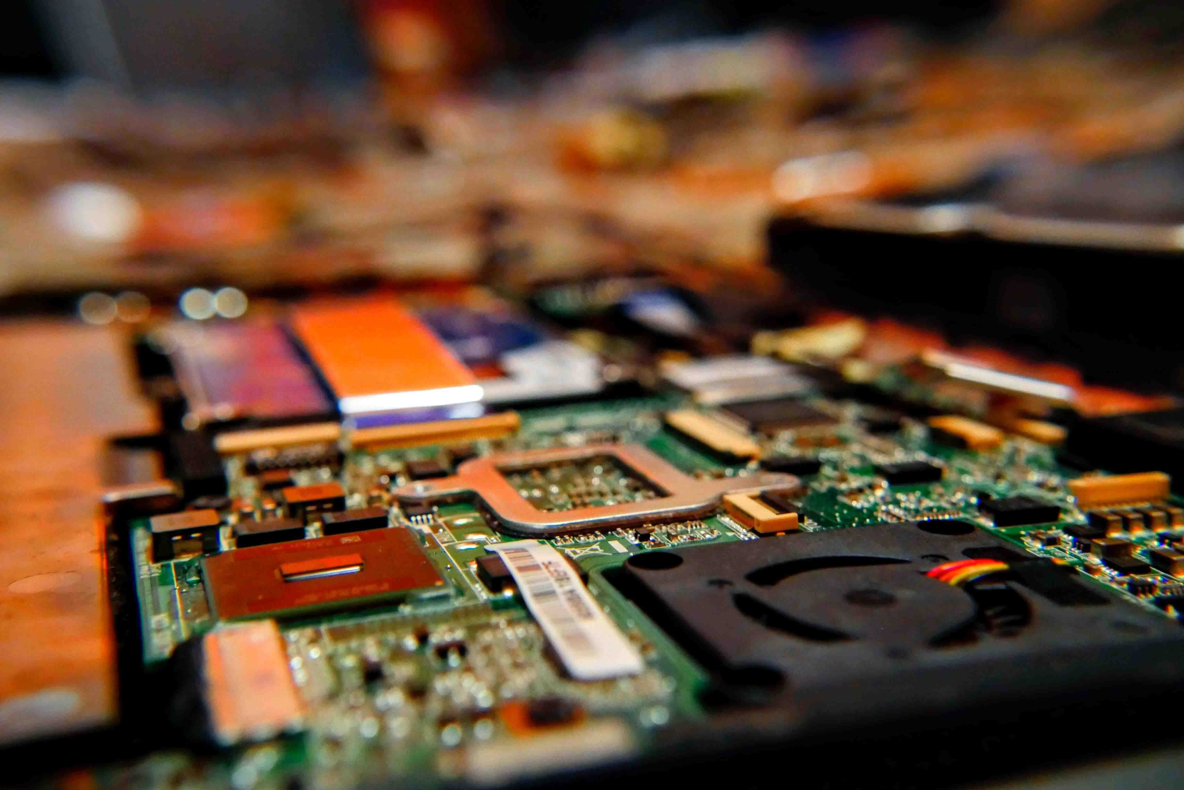 The image showcases a close-up view of a green computer motherboard on a wooden table. It features mounted electronic components, including a CPU chip, a heatsink or fan, and connected red and black wires. The setting suggests a technical atmosphere, possibly a workshop or laboratory dedicated to electronic device repair, maintenance, or study.